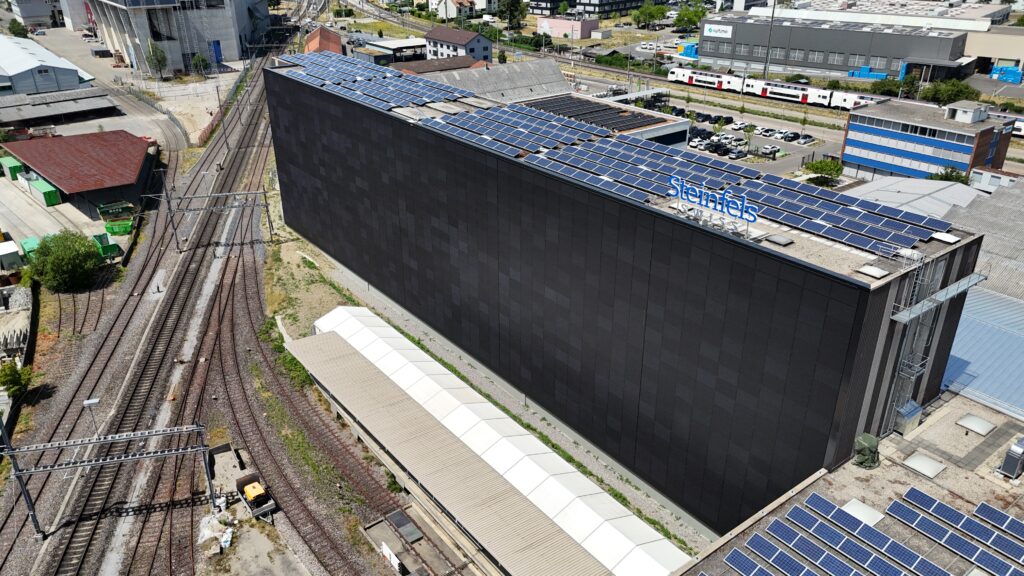 Aerial view of a dark building with solar panels on its roof beside a railway yard and parked cars nearby.