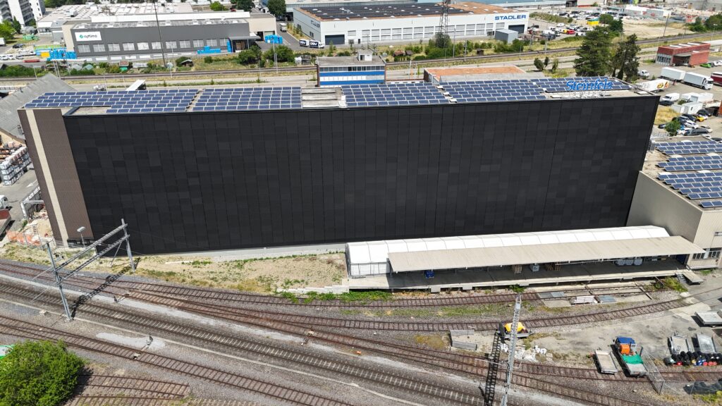 Aerial view of a large black industrial building with solar panels on its roof, beside train tracks in a busy industrial area.