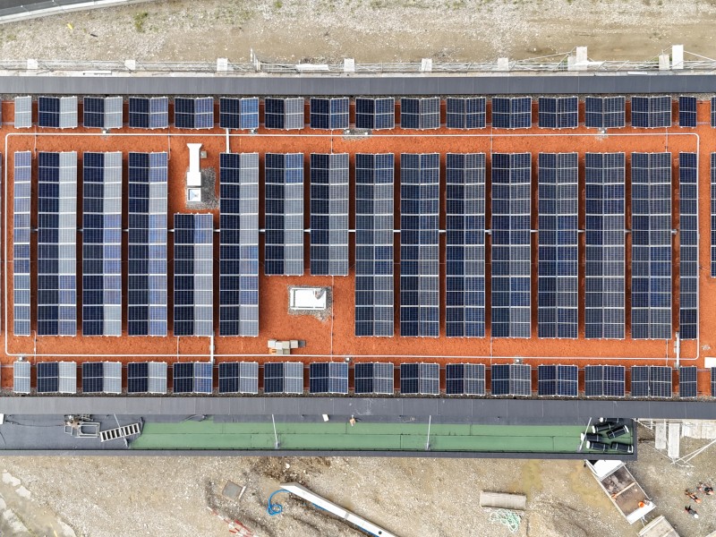 Aerial view of a solar panel farm on a flat red roof, with rows of blue panels and vents scattered across the roof surface.